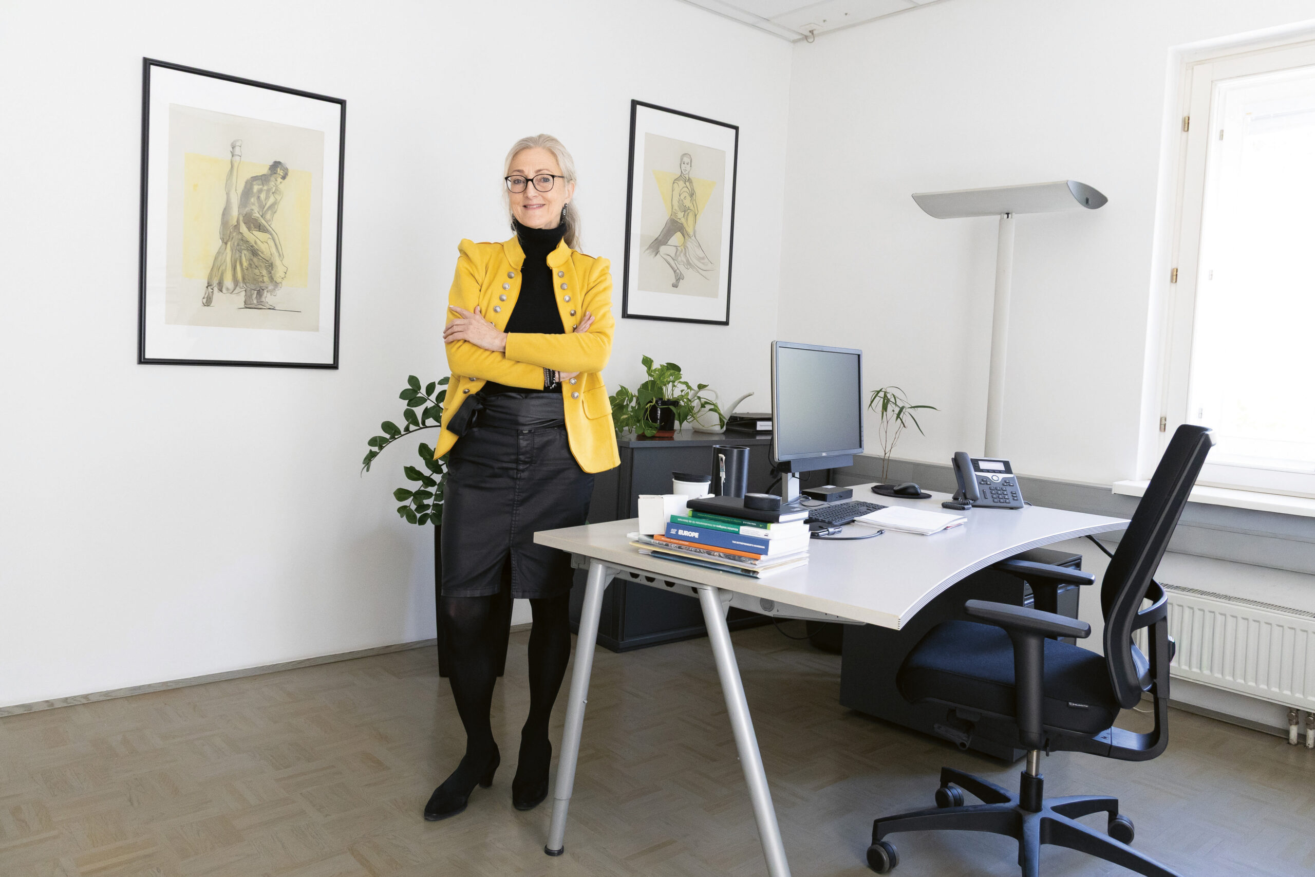 Eine Frau in einem gelben Blazer steht selbstbewusst an ihrem Schreibtisch, an dem Bücher, ein Computer und Pflanzen stehen; an den weißen Wänden hängt Kunst.