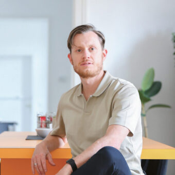 Zwei Männer sitzen auf schwarzen Stühlen in einem hellen, modernen Büro, der eine trägt ein dunkles Hemd, der andere ein helles Hemd, zwischen ihnen steht ein orangefarbener Tisch.