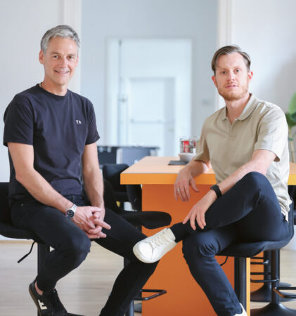 Zwei Männer sitzen auf hohen Stühlen neben einem orangefarbenen Tisch in einem hellen, modernen Raum mit Holzboden und weißen Wänden und schauen in die Kamera.