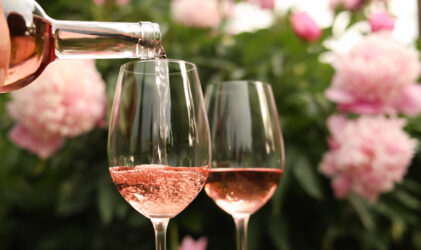 Roséwein wird in ein Glas gegossen, daneben steht ein weiteres gefülltes Glas vor einem Hintergrund aus rosa Blumen und Grünzeug.