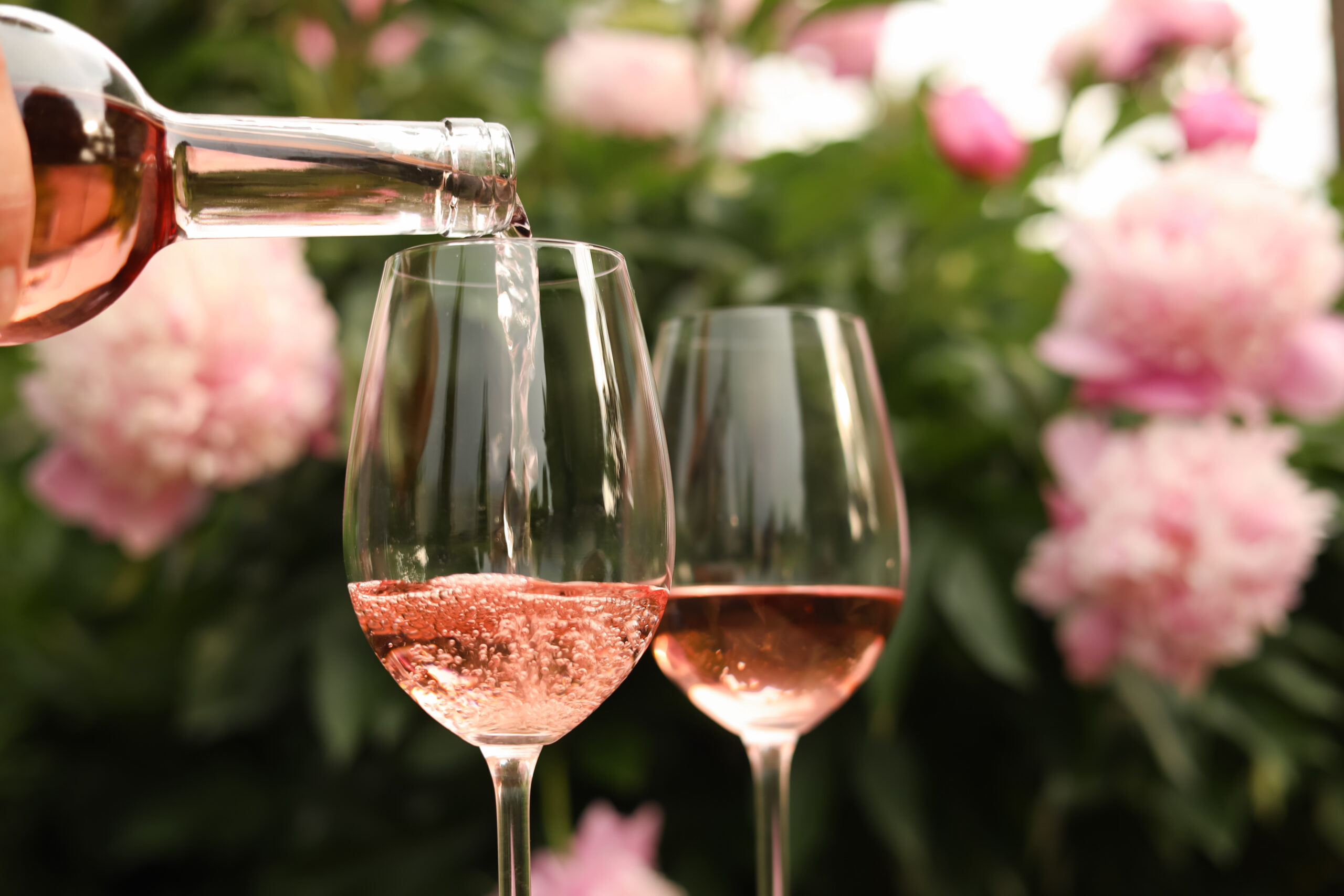 Roséwein wird in ein Glas gegossen, daneben steht ein weiteres gefülltes Glas vor einem Hintergrund aus rosa Blumen und Grünzeug.