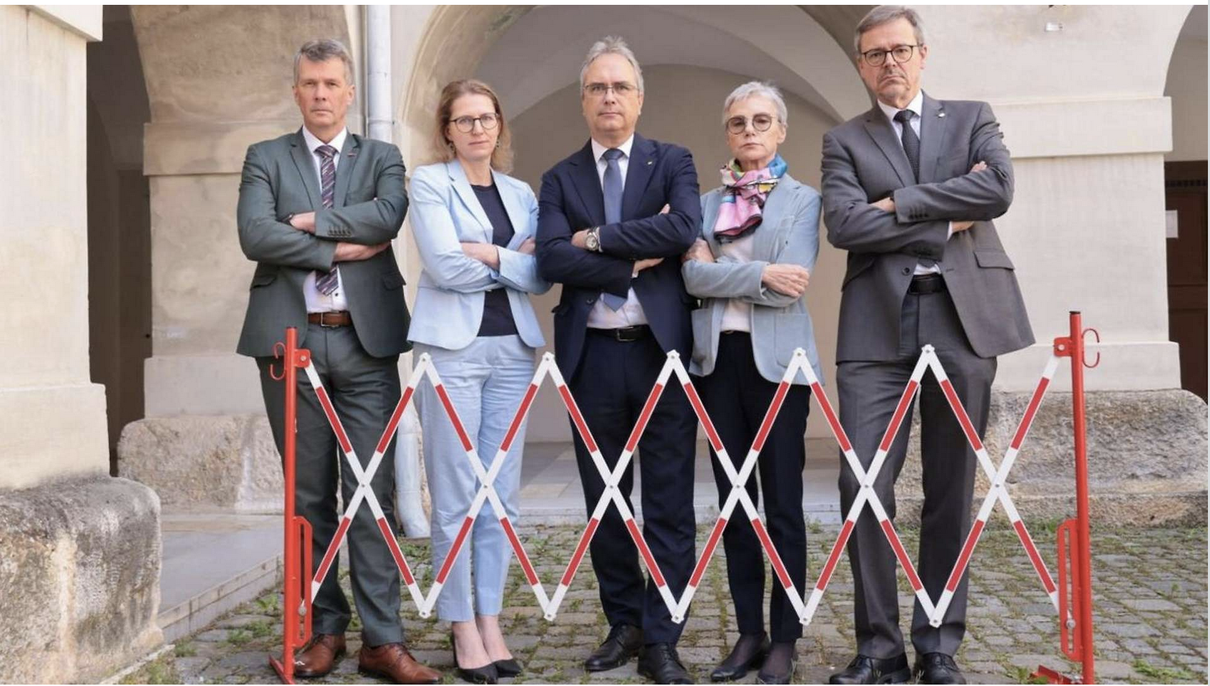 Fünf Erwachsene in Geschäftskleidung stehen mit verschränkten Armen hinter einer Schranke der GRAWE Bilanz 2025 und posieren vor einer gewölbten Gebäudehülle.
