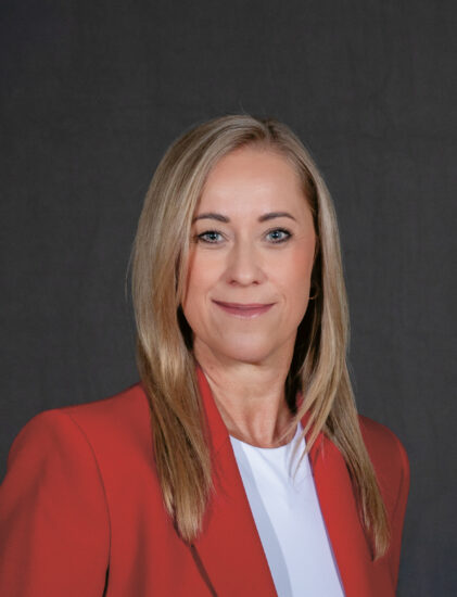 Eine Frau mit glattem blondem Haar, die einen roten Blazer und ein weißes Oberteil trägt, posiert vor einem dunkelgrauen Hintergrund.