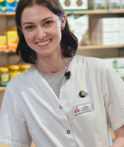 Eine lächelnde Frau in einem weißen Apothekenkittel steht vor Gesundheitsprodukten.