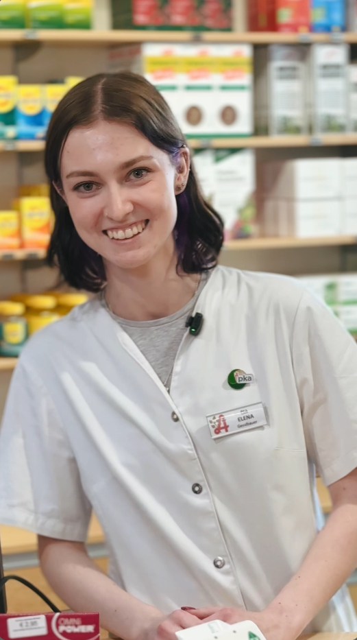 Eine lächelnde Frau in einem weißen Apothekenkittel steht vor Gesundheitsprodukten.