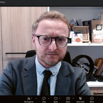 Ein Mann im Anzug und mit Brille sitzt während einer Zoom-Sitzung in einem Büro, hinter ihm stehen Bücher und Auszeichnungen in Regalen.