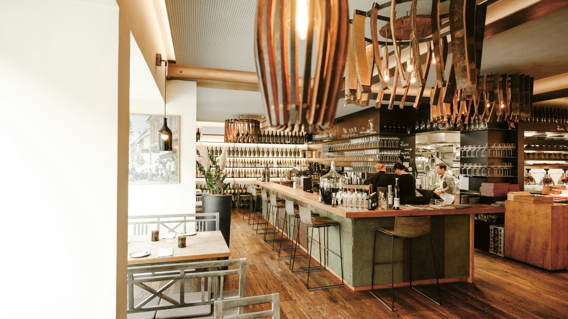 Modernes Restaurantinterieur mit Holzböden, Hängeleuchten, einem Barbereich und mit Flaschen gefüllten Regalen; das Personal arbeitet hinter der Bar.