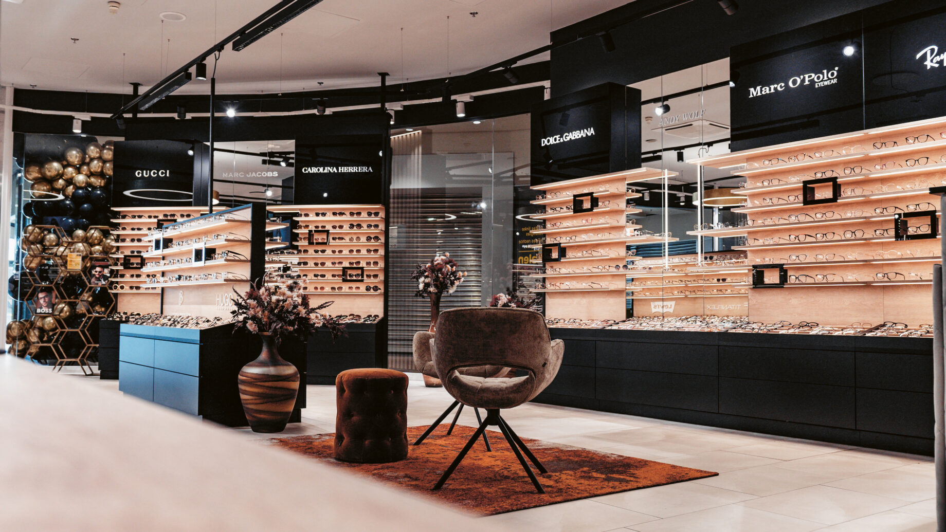 Modernes Brillengeschäft mit stilvollen Stühlen, dekorativen Pflanzen und Auslagen von Designer-Brillenmarken in Holzregalen.