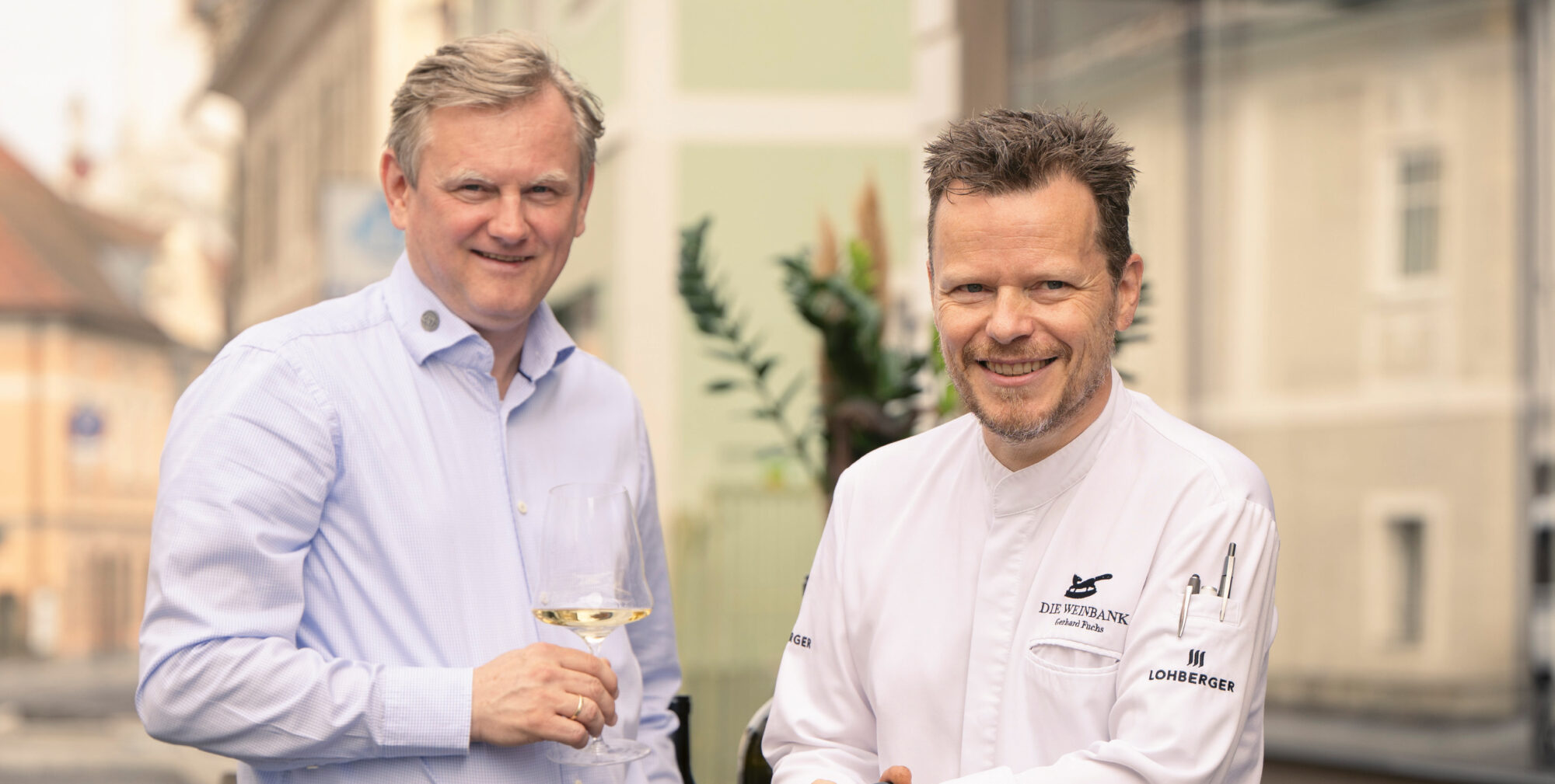Zwei Männer stehen vor einem Restaurant, einer mit einem Weinglas in der Hand, der andere im Kochmantel, vor ihnen Weinflaschen und Kisten mit der Aufschrift "Tement".