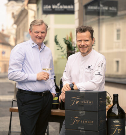 Zwei Männer stehen vor einem Restaurant, einer mit einem Weinglas in der Hand, der andere im Kochmantel, vor ihnen Weinflaschen und Kisten mit der Aufschrift "Tement".