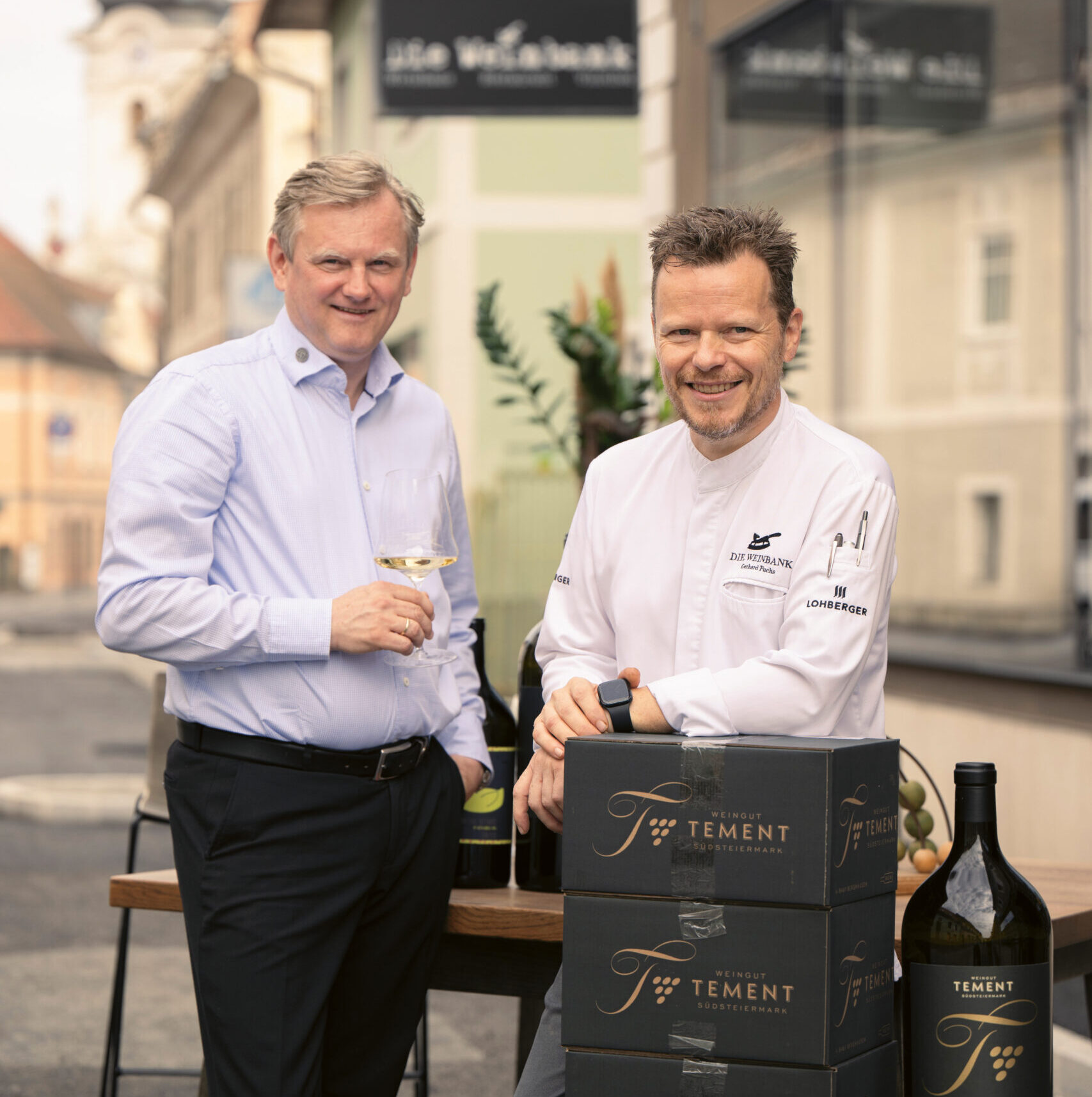 Zwei Männer stehen vor einem Restaurant, einer mit einem Weinglas in der Hand, der andere im Kochmantel, vor ihnen Weinflaschen und Kisten mit der Aufschrift "Tement".