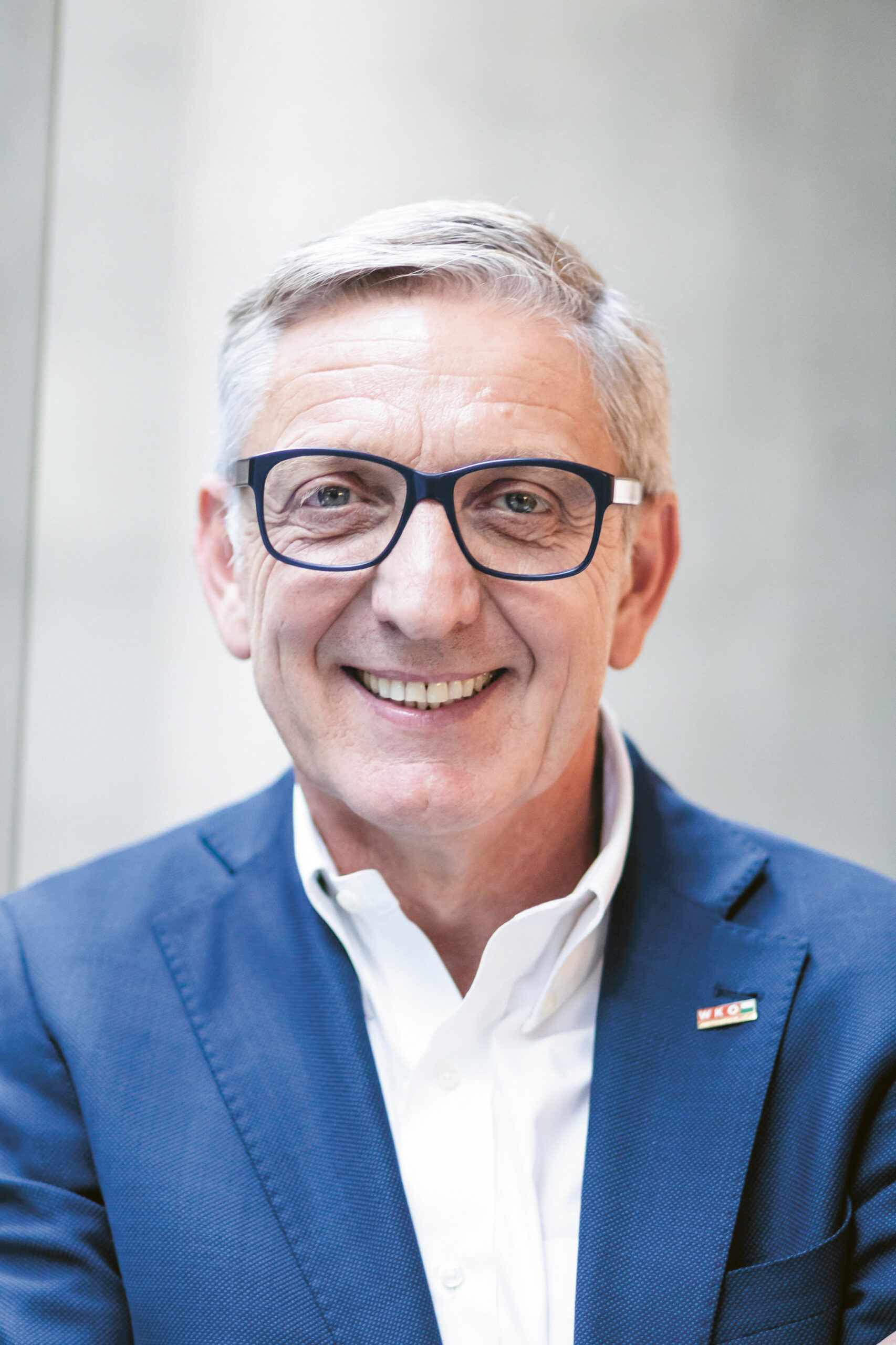 Ein lächelnder älterer Mann mit Brille und blauem Sakko, an dem eine Anstecknadel steckt, steht vor einem hellen, unscharfen Hintergrund.