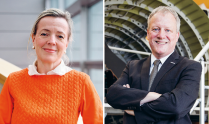 Eine Frau in einem orangefarbenen Pullover und ein Mann im Anzug stehen getrennt voneinander und lächeln beide in einem Innenraum vor einem industriellen und modernen Hintergrund.