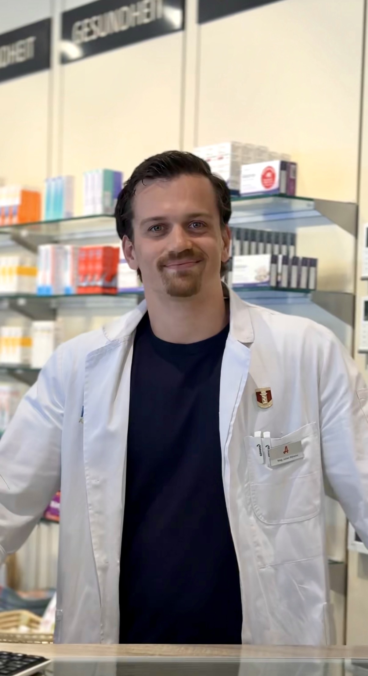 Eine lächelnde Apotheker, Julian Bleierer steht vor mehreren Apothekenprodukten hinter der Tara.