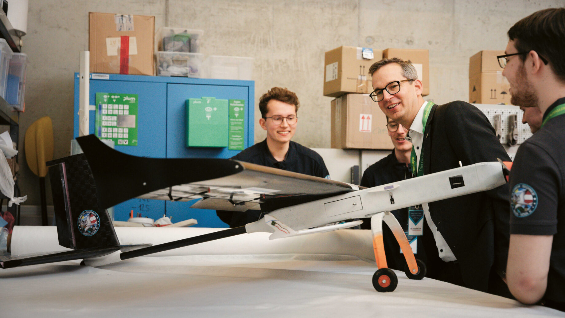 Vier Menschen stehen lächelnd um ein großes Flugzeugmodell herum in einer Werkstatt, die mit Regalen, Kisten und Werkzeugen gefüllt ist.