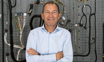 Ein Mann in einem hellblauen Hemd steht lächelnd mit verschränkten Armen vor einer an der Wand befestigten Tafel, auf der bunte Elektrokabel und -drähte zu sehen sind.