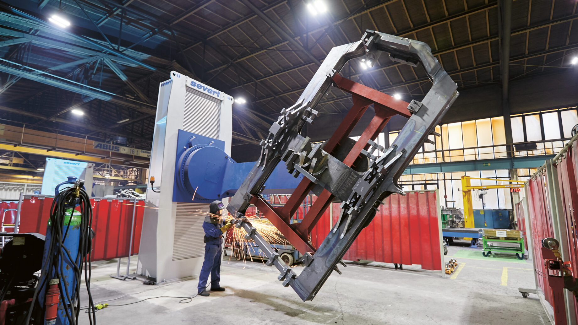 Ein Arbeiter in einer Fabrik schweißt mit Funkenflug einen großen Metallrahmen, wobei er in einer geräumigen, hell erleuchteten Werkstatt Industriemaschinen einsetzt.