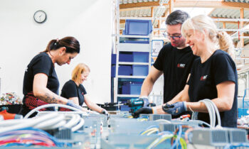 Vier Personen in schwarzen Hemden arbeiten gemeinsam an einem Arbeitsplatz in einer hellen Industrieumgebung mit freiliegenden Balken daran, elektronische Bauteile zusammenzubauen.