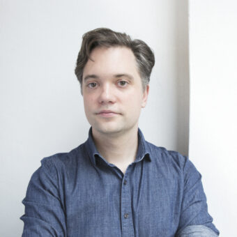 Ein Mann mit heller Haut und kurzen braunen Haaren steht an einer weißen Wand. Er trägt ein blaues Button-up-Hemd mit hochgekrempelten Ärmeln und verschränkten Armen.