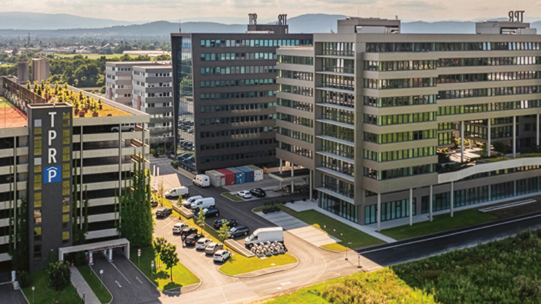 Luftaufnahme moderner Bürogebäude mit begrünten Dächern, Parkplätzen und Grünflächen im Vordergrund unter strahlend blauem Himmel.