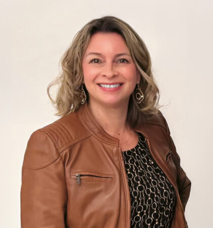Lächelnde Frau mit welligem blondem Haar, die eine braune Lederjacke über einem gemusterten schwarzen Kleid trägt und vor einem schlichten hellen Hintergrund steht.
