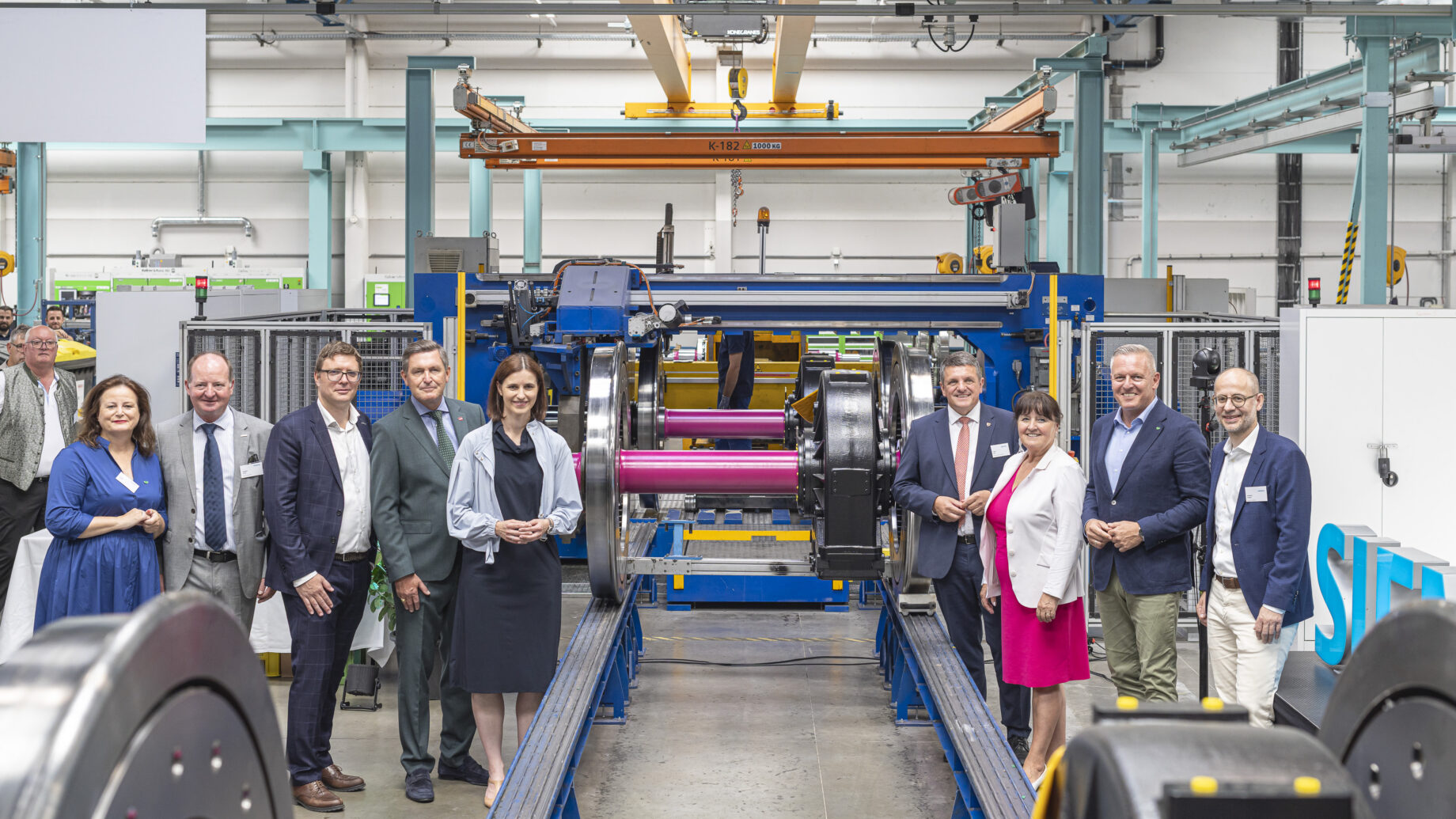Eine Gruppe in Geschäftskleidung lächelt vor Industriemaschinen und feiert „125 Jahre Siemens in der Steiermark“ in einer hellen, modernen Fabrikhalle.