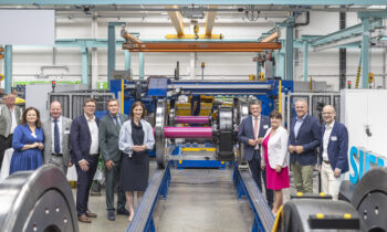 Eine Gruppe in Geschäftskleidung lächelt vor Industriemaschinen und feiert „125 Jahre Siemens in der Steiermark“ in einer hellen, modernen Fabrikhalle.
