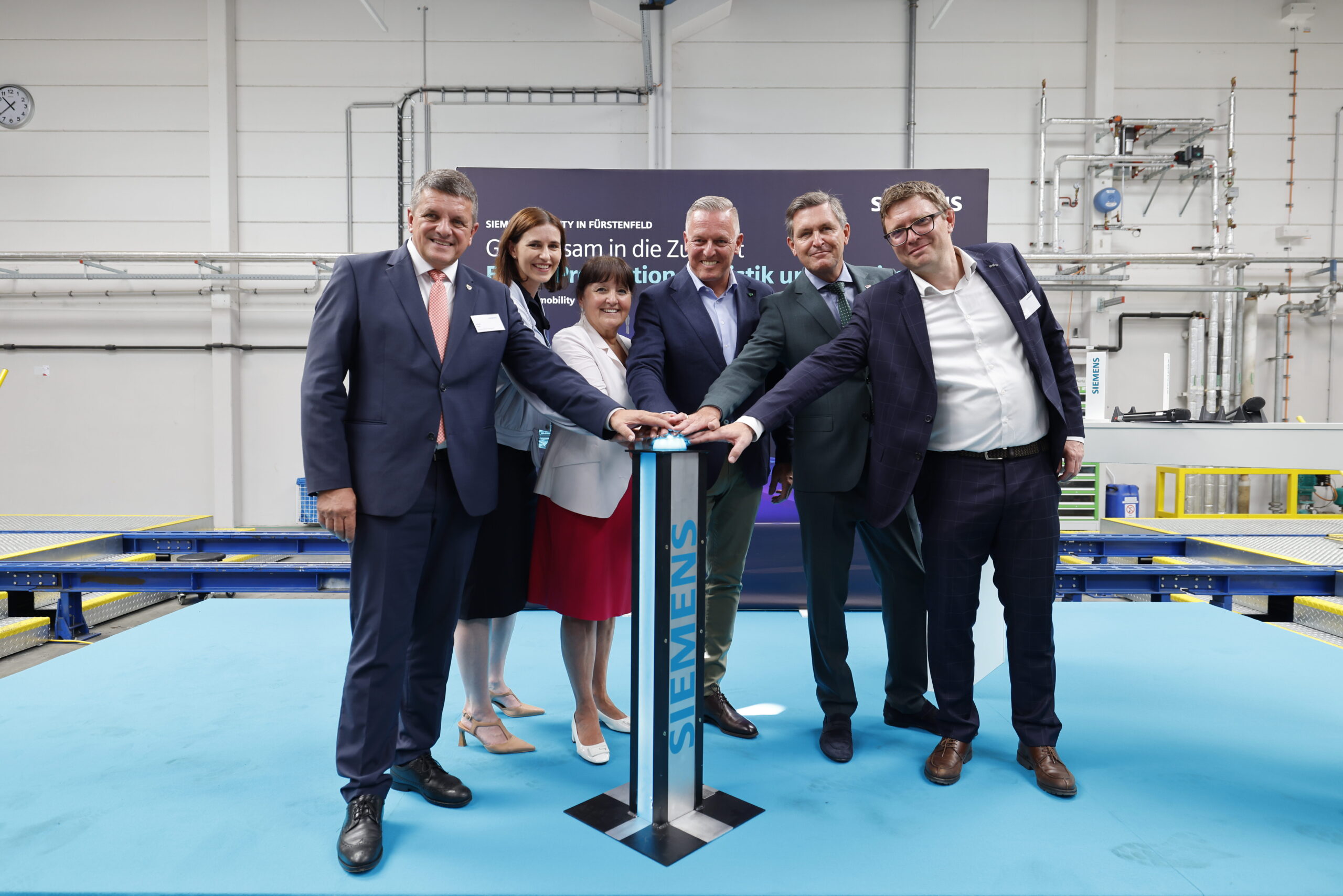 Sechs Personen in Geschäftskleidung feiern „125 Jahre Siemens in der Steiermark“ – lächelnd drücken sie in einer industriellen Umgebung einen Knopf auf einem Podium.