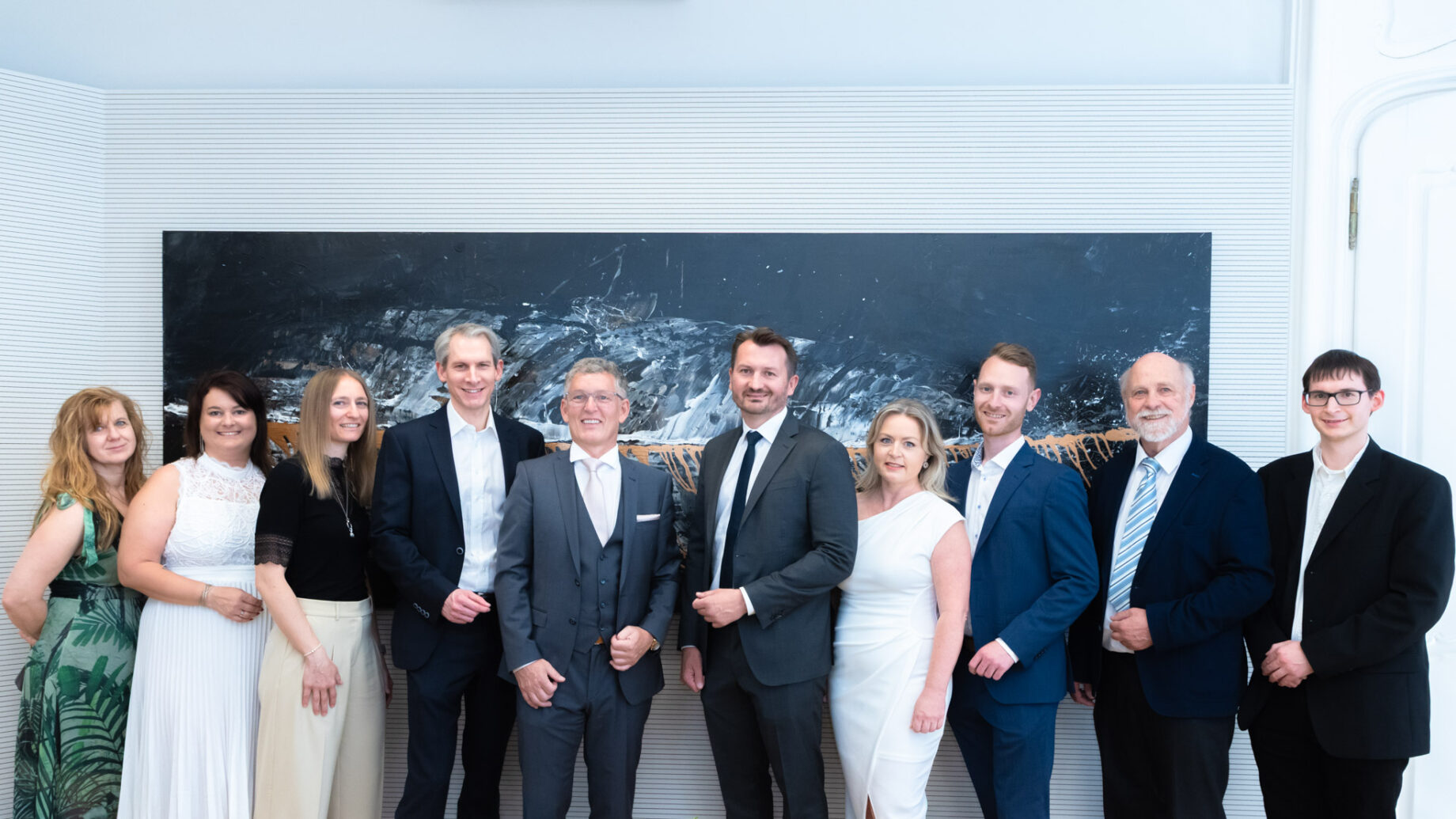 Eine Gruppe von zehn Personen, förmlich gekleidet in Anzügen und Kleidern, steht lächelnd vor einem großen, dunklen Kunstwerk an einer weißen Wand.