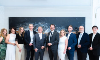 Eine Gruppe von zehn Personen, förmlich gekleidet in Anzügen und Kleidern, steht lächelnd vor einem großen, dunklen Kunstwerk an einer weißen Wand.