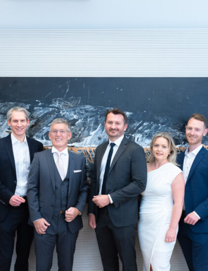 Eine Gruppe von zehn Personen, förmlich gekleidet in Anzügen und Kleidern, steht lächelnd vor einem großen, dunklen Kunstwerk an einer weißen Wand.
