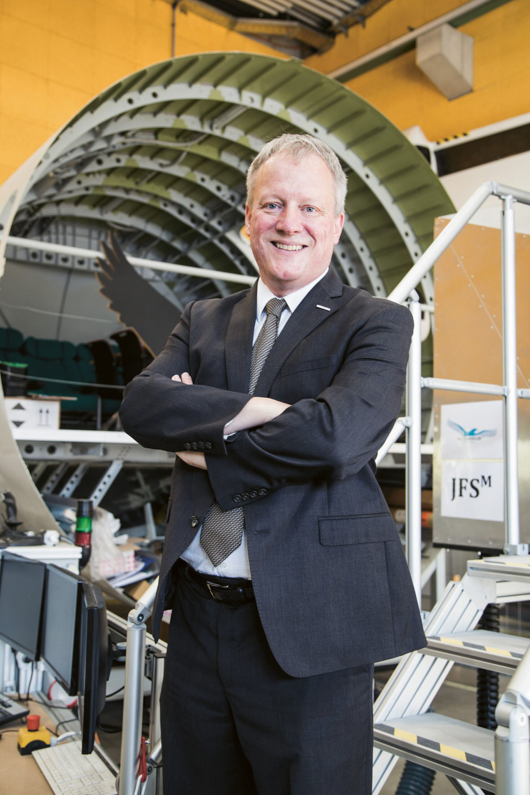 Ein lächelnder Mann im Anzug steht mit verschränkten Armen vor einem großen, runden Industriegebäude und Bürogeräten.