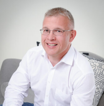 Ein lächelnder Mann mit Brille, weißem Hemd und Jeans sitzt auf einem grauen Sessel mit einem gemusterten Kissen, die Hände ineinander verschränkt.