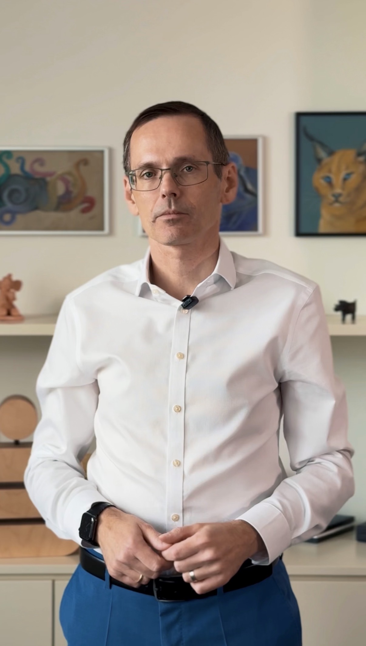 Ein Mann mit Brille und weißem Hemd steht in einem Innenraum, hinter ihm Regale und ein Katzenbild