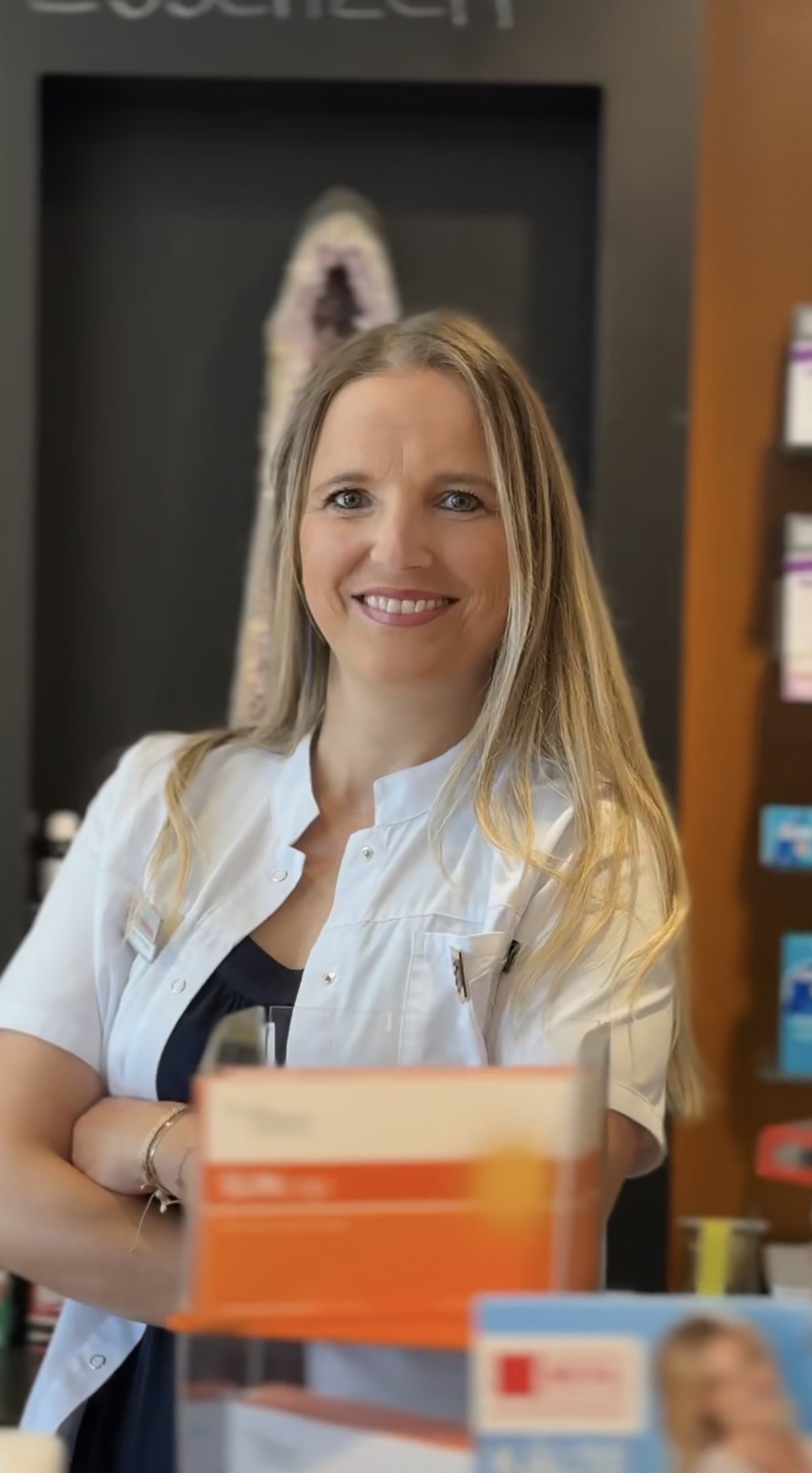 Eine lächelnde Frau in einem weißen Apothekenkittel steht mit verschränkten Armen da.