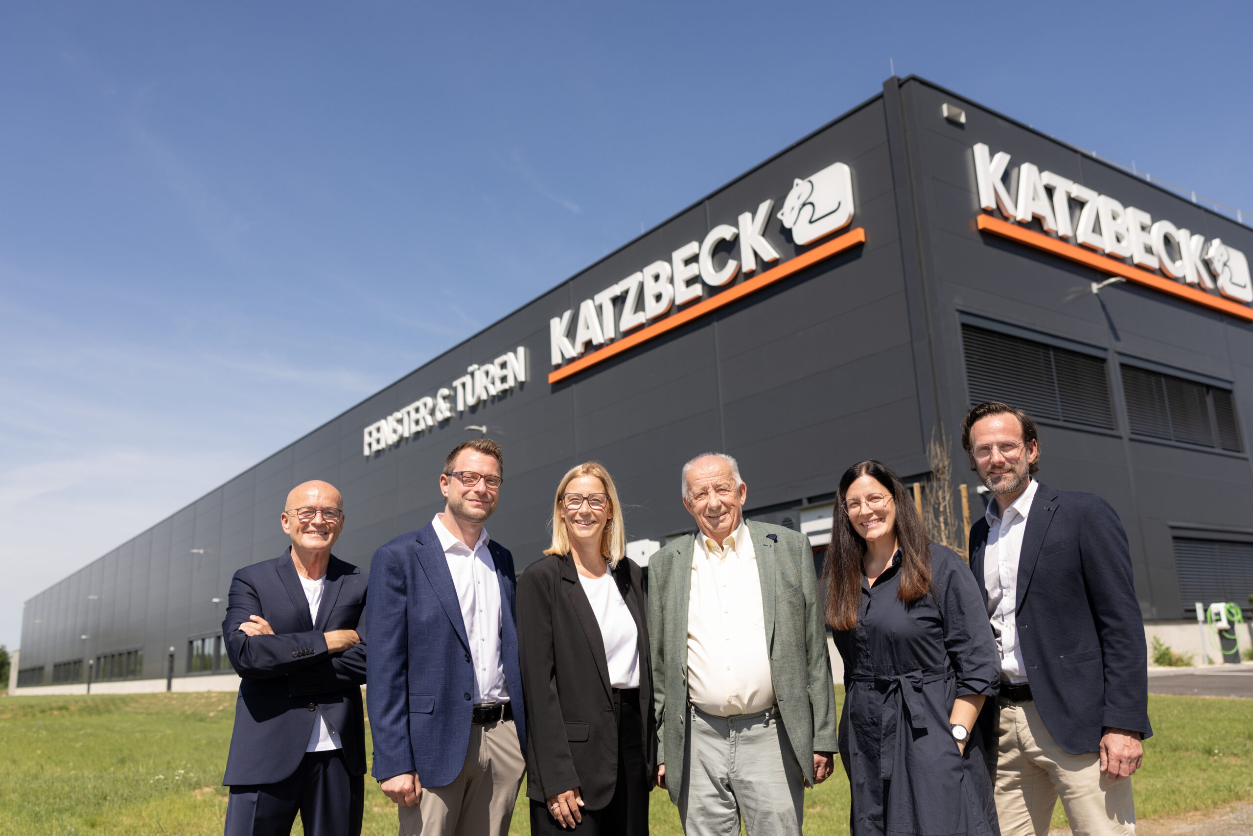 Sechs Personen in Geschäftskleidung lächeln an einem sonnigen Tag vor dem modernen Katzbeck-Gebäude - dem Sitz von Katzbeck Fenster, Testsieger bei Markentreue.