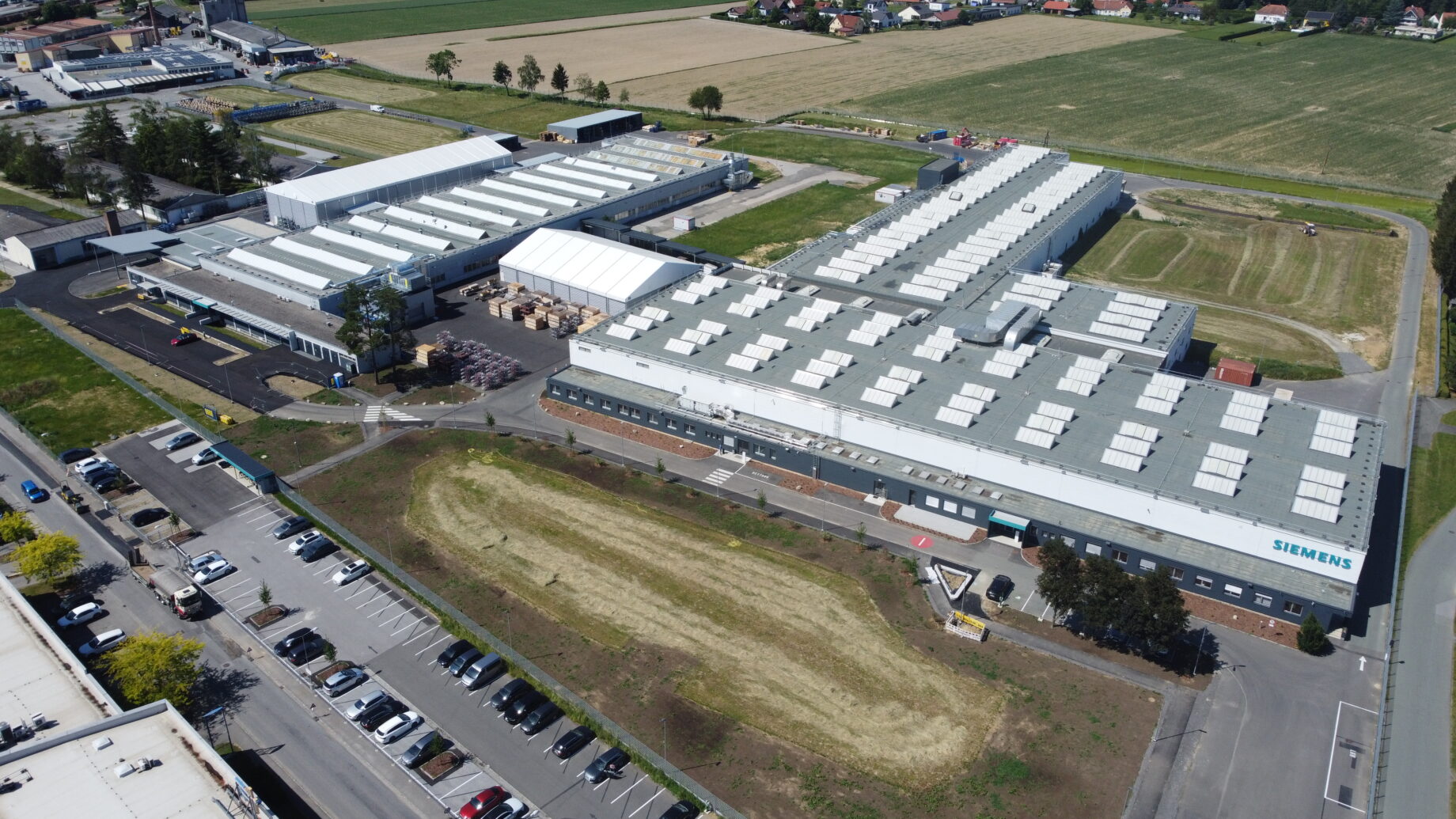 Luftaufnahme eines großen Siemens-Werks anlässlich des Jubiläums „125 Jahre Siemens in der Steiermark“, umgeben von Parkplätzen, Feldern und Straßen.