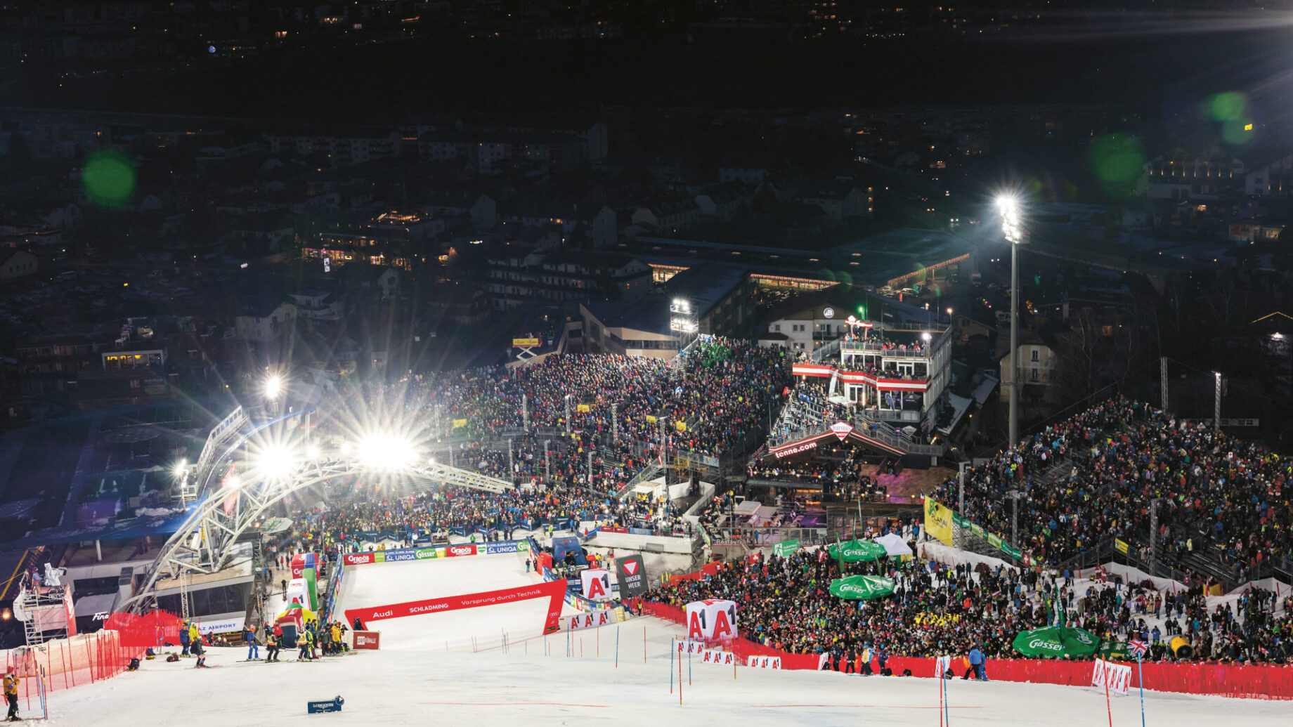 Nachts versammelt sich eine große Menschenmenge in der Nähe einer hell beleuchteten Skipiste, im Hintergrund sind Tribünen und Gebäude zu sehen.