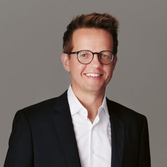 Ein lächelnder Mann mit kurzen braunen Haaren und Brille, der ein dunkles Sakko über einem weißen Hemd trägt und vor einem schlichten grauen Hintergrund steht.