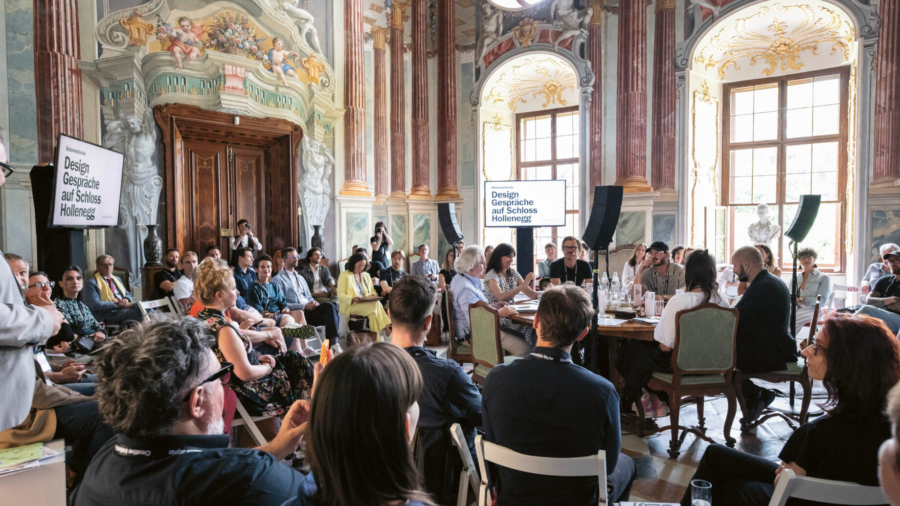 Eine große Gruppe von Menschen sitzt in einem prächtigen, verzierten Raum mit hohen Decken und Wandmalereien und nimmt an einer Designkonferenz oder einer Diskussion an Tischen teil.
