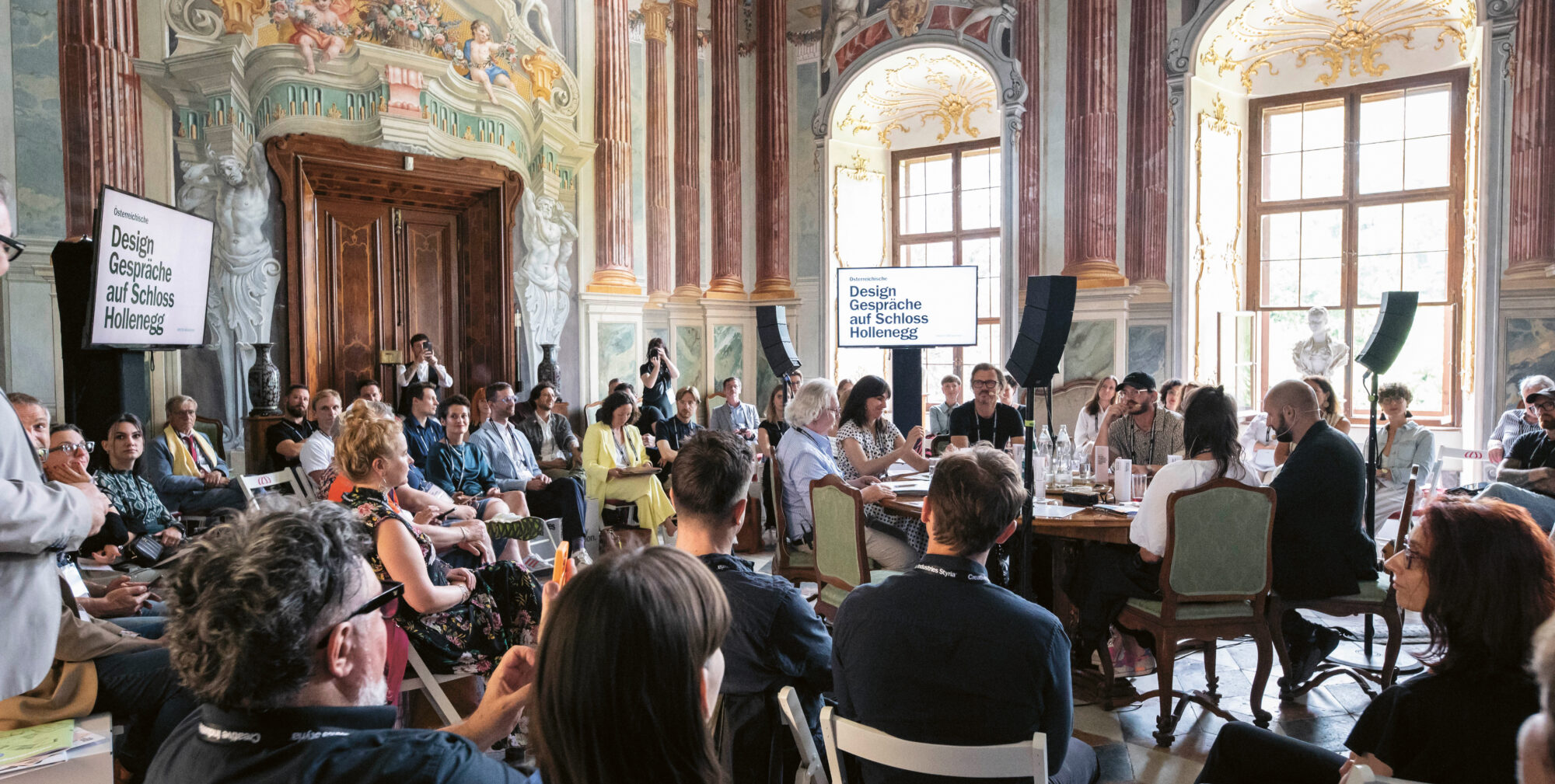 Eine große Gruppe von Menschen sitzt in einem prächtigen, verzierten Raum mit hohen Decken und Wandmalereien und nimmt an einer Designkonferenz oder einer Diskussion an Tischen teil.