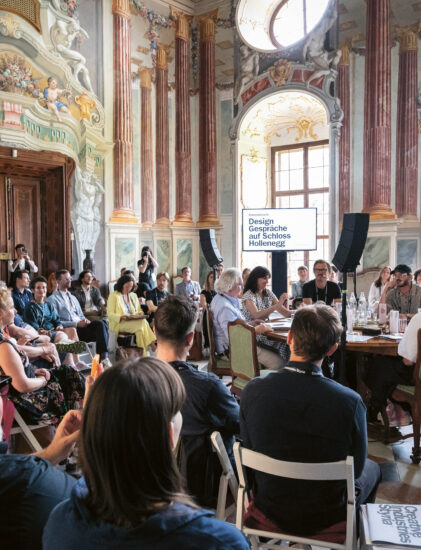 Eine große Gruppe von Menschen sitzt in einem prächtigen, verzierten Raum mit hohen Decken und Wandmalereien und nimmt an einer Designkonferenz oder einer Diskussion an Tischen teil.