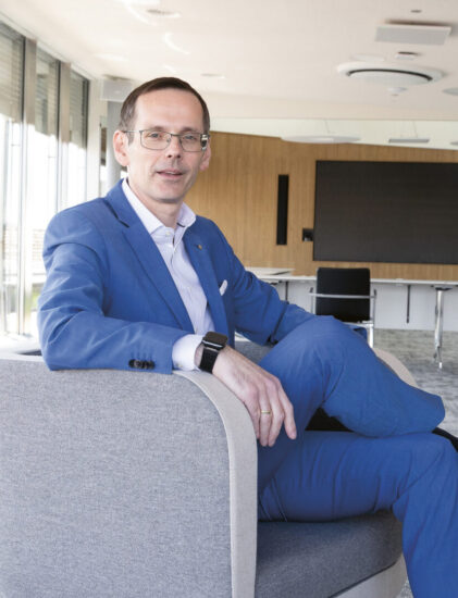Ein Mann in einem blauen Anzug sitzt mit überkreuzten Beinen auf einem modernen Bürostuhl, im Hintergrund ist ein großer, heller Konferenzraum zu sehen.