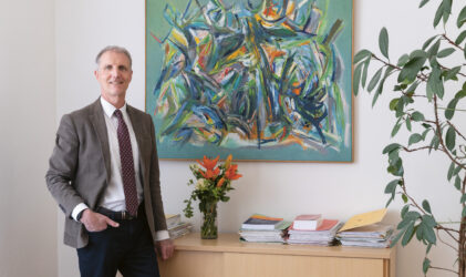 Ein Mann im Anzug steht neben einem Schrank, auf dem Blumen, Bücher und Papiere liegen; im Hintergrund sind abstrakte Kunstwerke und eine Pflanze zu sehen.