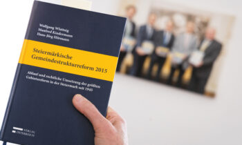 Eine Hand hält ein Buch mit dem Titel „Steiermärkische Gemeindestrukturreform 2015“, im Hintergrund sind verschwommene Menschen zu sehen.
