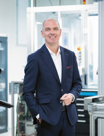 Ein lächelnder Mann in einem blauen Anzug steht in einer modernen, hellen Industrie- oder Laborumgebung mit verschiedenen Geräten und Maschinen im Hintergrund.