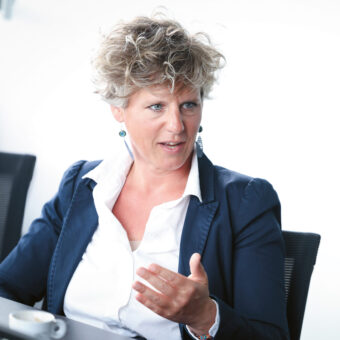 Eine Frau mit lockigem Haar in einem marineblauen Blazer gestikuliert, während sie bei einer Besprechung spricht; vor ihr auf dem Tisch steht eine Kaffeetasse.
