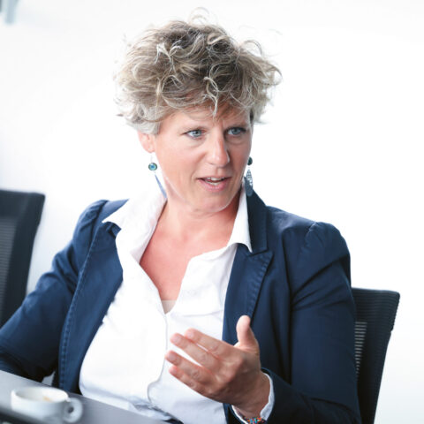 Eine Frau mit lockigem Haar in einem marineblauen Blazer gestikuliert, während sie bei einer Besprechung spricht; vor ihr auf dem Tisch steht eine Kaffeetasse.