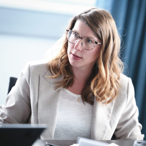 Eine Frau mit welligem, hellbraunem Haar und Brille sitzt an einem Tisch, wirkt aufmerksam und ist in ein Gespräch vertieft. Sie trägt einen beigen Blazer und ein weißes Oberteil.