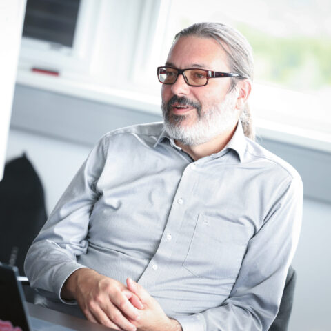 Ein Mann mit Brille und grauem Hemd sitzt lächelnd an einem Schreibtisch, im Hintergrund sind ein Laptop und ein Whiteboard zu sehen.